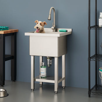 A small dog sits inside a stainless steel utility sink equipped with a high-arc faucet in a modern, gray-walled grooming area. The scene includes a wooden-top workbench, a wire shelving unit stocked with pet supplies, and a set of metal food bowls on the floor.