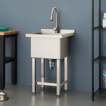 A sleek, stainless steel utility sink with a high-arc faucet stands against a slate blue wall in a modern laundry or utility room. The space is organized with a wooden workbench to the left and a black wire shelving unit holding various household supplies to the right.