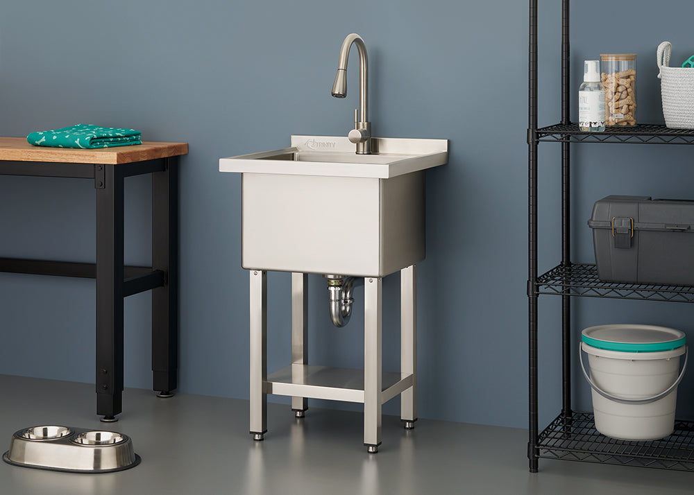 A sleek, stainless steel utility sink with a high-arc faucet stands against a slate blue wall in a modern laundry or utility room. The space is organized with a wooden workbench to the left and a black wire shelving unit holding various household supplies to the right.