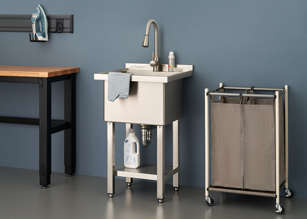 A stainless steel utility sink with a tall gooseneck faucet stands against a blue wall between a wooden workbench and a rolling laundry sorter. The sink features a bottom storage shelf holding a bottle of detergent, with a gray cloth draped over the basin's edge.