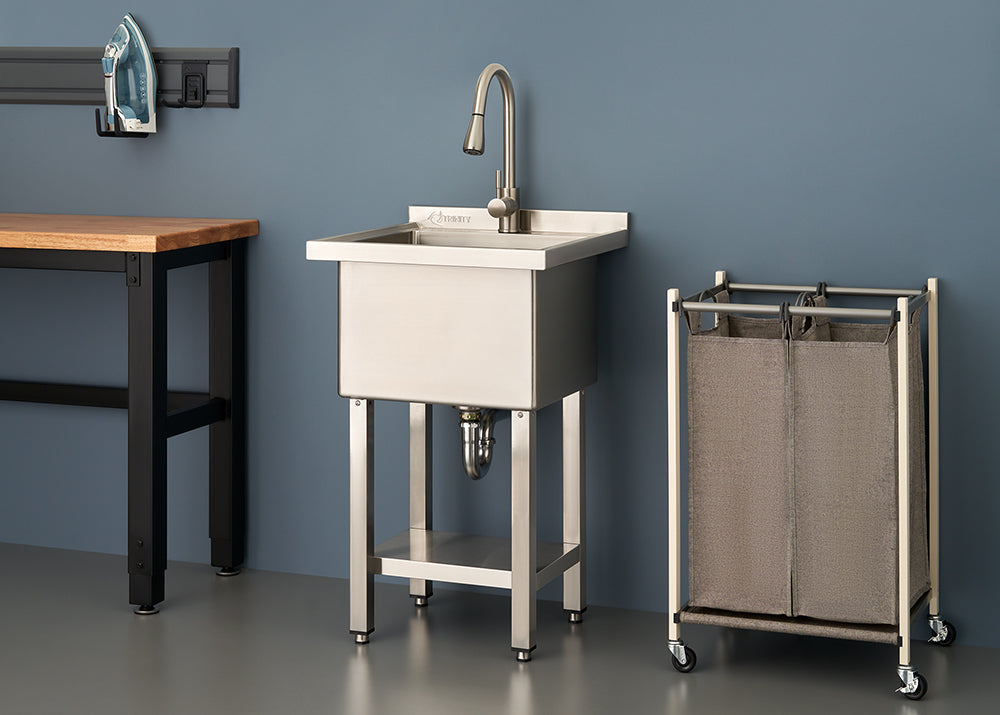 A stainless steel utility sink with a tall gooseneck faucet stands in a modern laundry room or workshop between a wooden workbench and a rolling laundry sorter. The scene is set against a dark blue wall and features clean, industrial-style organization.
