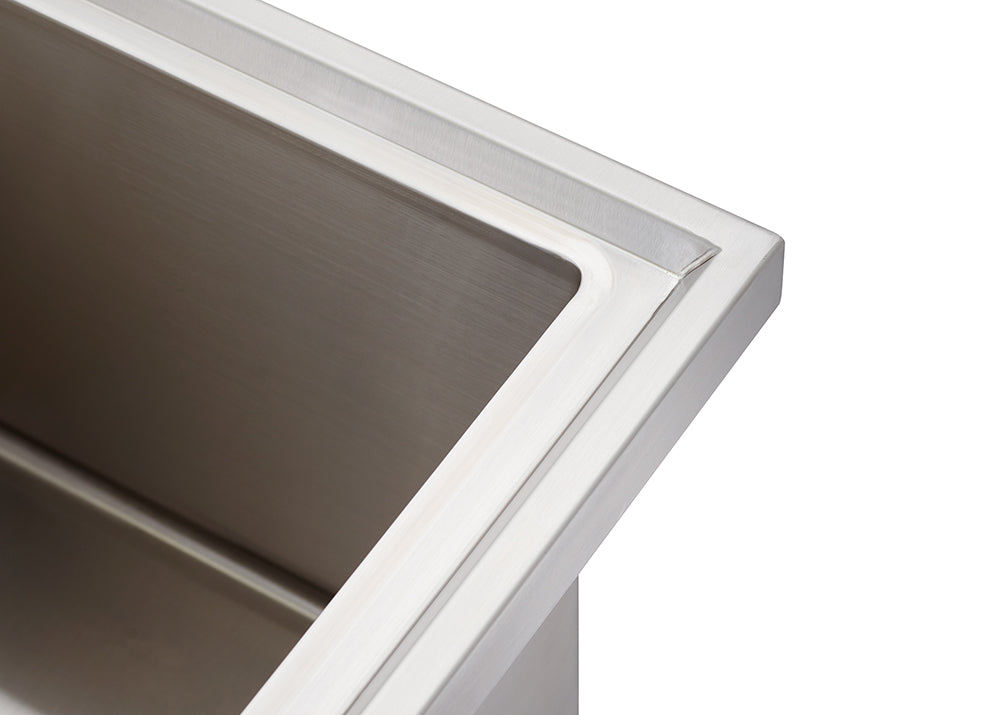 A close-up shot shows the corner of a brushed stainless steel rectangular sink with a wide, flat rim. The image highlights the clean lines and metallic texture of the sink's basin and mounting edge.