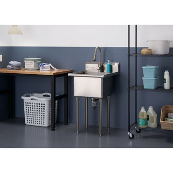 A stainless steel utility sink with a gooseneck faucet stands in a laundry or utility room. To the left is a wooden table with various cleaning and laundry supplies, and to the right is a black wire shelving unit holding more supplies.