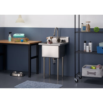 A small, fluffy, light brown and white dog stands in a stainless steel utility sink with a high-arc pull-out faucet. The sink is positioned in a room with a two-toned blue and white wall, a wooden table with pet grooming supplies, a rolling metal shelf unit with various items, and a gray rug on the floor.