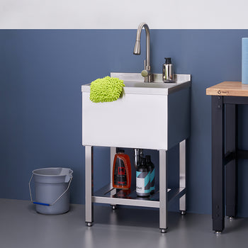 A close-up shot shows a stainless steel utility sink with a tall faucet, a green scrubbing mitt, and two bottles of cleaning solution on the bottom shelf. To the left is a grey bucket, and to the right, a wooden-topped workbench holds two rolls of blue paper towels.