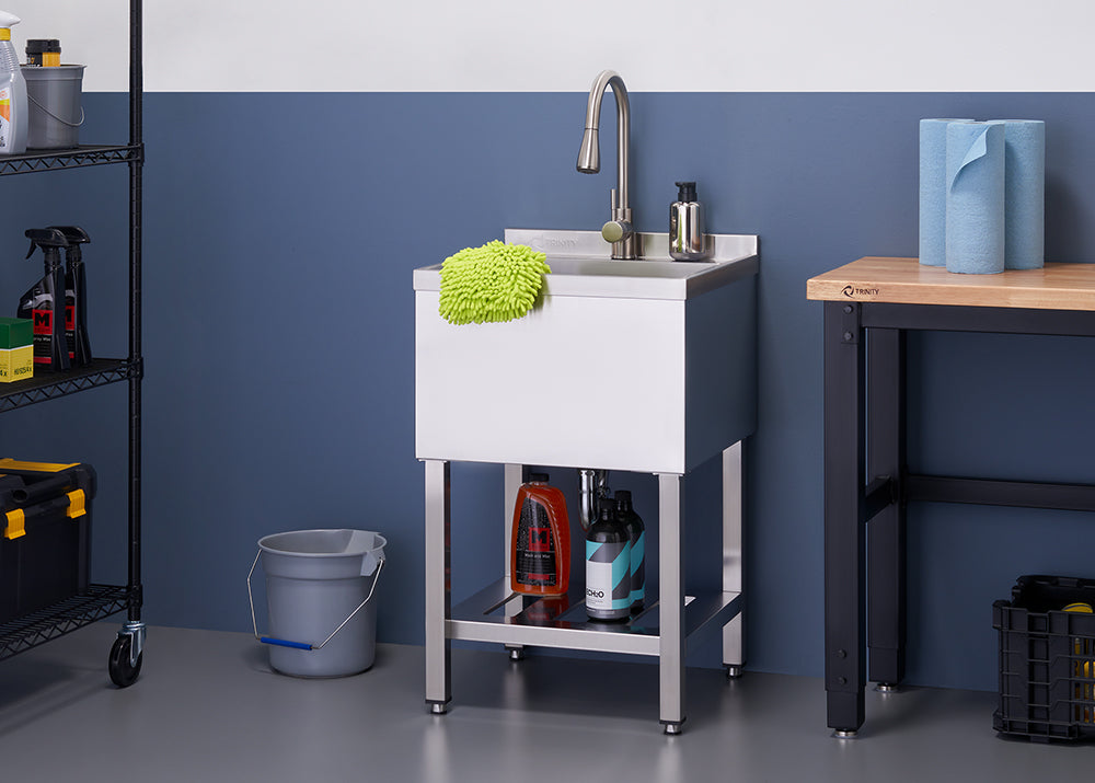 A close-up shot shows a stainless steel utility sink with a tall faucet, a green scrubbing mitt, and two bottles of cleaning solution on the bottom shelf. To the left is a grey bucket, and to the right, a wooden-topped workbench holds two rolls of blue paper towels.
