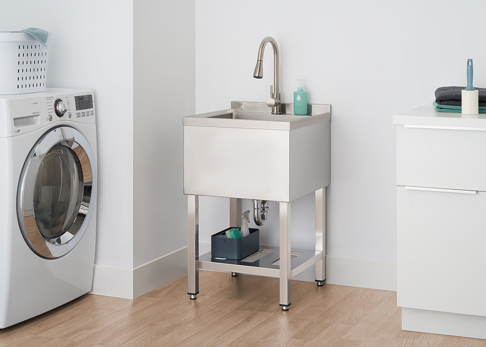 A polished stainless steel utility sink with a gooseneck faucet stands in a laundry room. Cleaning supplies are stored on a shelf beneath the sink, next to a white washing machine.