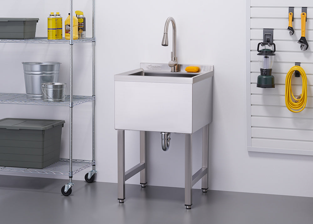 A chrome utility sink with a faucet stands against a white wall in a well-organized garage or workspace. To the left, a metal shelving unit holds various items including buckets and storage bins, while tools and a yellow extension cord hang on a pegboard to the right.