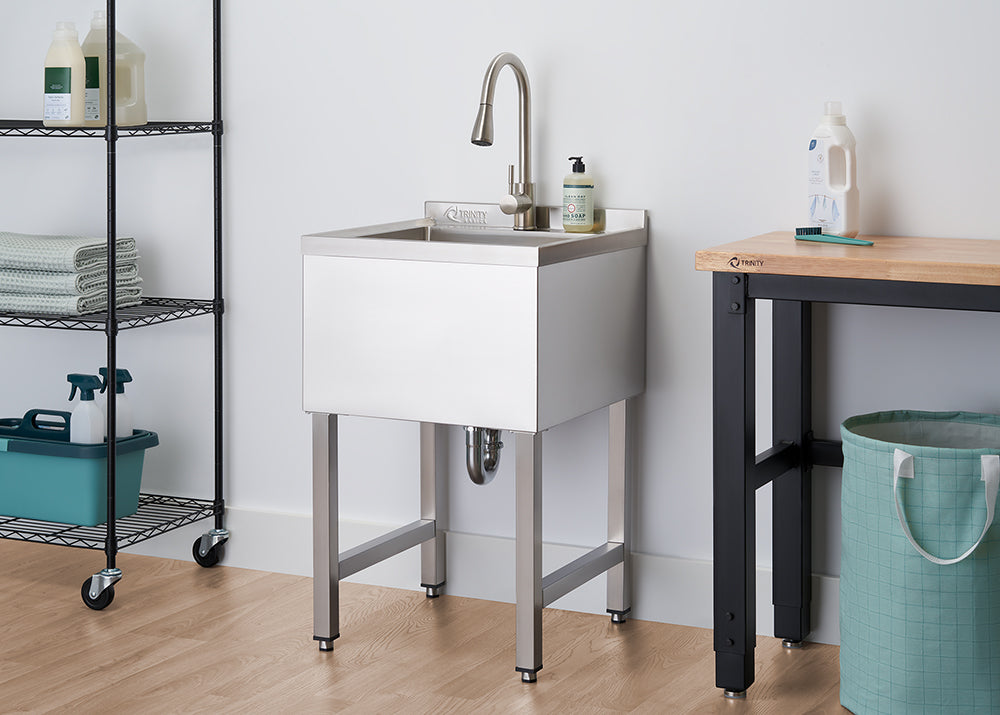 A chrome utility sink with a faucet stands against a white wall in a laundry room. To its left, a black wire shelving unit on wheels holds towels and cleaning supplies, while a wooden workbench with black metal legs is visible to its right.