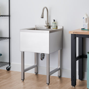 A chrome utility sink with a faucet stands against a white wall in a laundry room. To its left, a black wire shelving unit on wheels holds towels and cleaning supplies, while a wooden workbench with black metal legs is visible to its right.