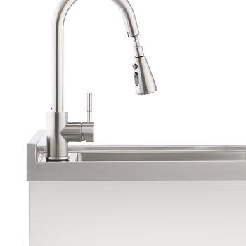 A close-up shot shows a brushed stainless steel pull-down faucet with a single handle, installed on a stainless steel utility sink. The faucet's sprayer head is slightly angled downwards, indicating its pull-down feature.