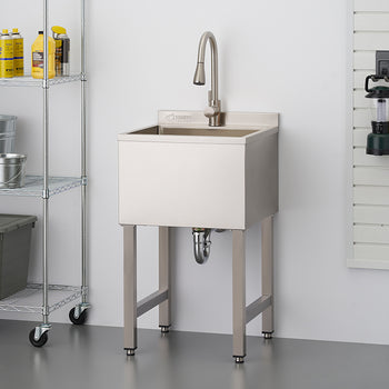 A stainless steel utility sink with a high-arc faucet stands in a garage or workshop setting next to a wire shelving unit and a tool slatwall. The freestanding sink features a deep basin and adjustable leveling feet on its metal legs.