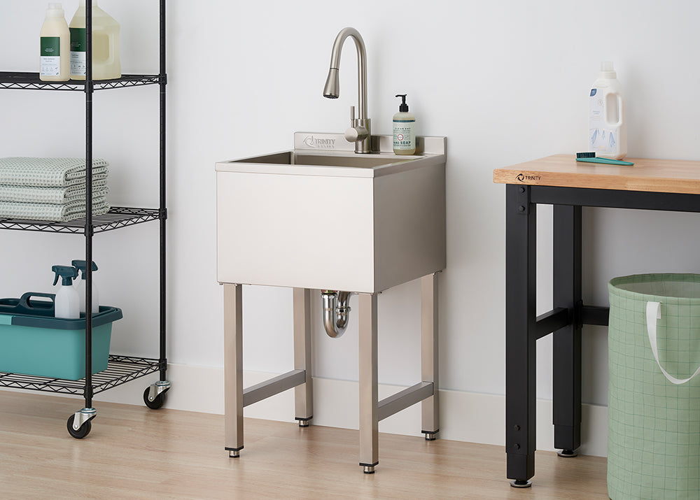 A modern stainless steel utility sink with a tall gooseneck faucet stands in a bright, organized laundry room between a wire shelving unit and a wooden workbench. The sink features a deep basin and square legs, with a visible chrome P-trap underneath.