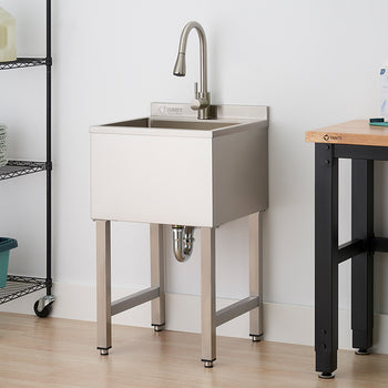 A stainless steel utility sink with a tall, arched faucet stands in a laundry room between a wire shelving rack and a wooden workbench. The sink is supported by four square metal legs, with the plumbing P-trap visible underneath against a white wall and light wood flooring.