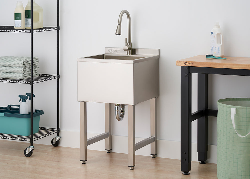 A stainless steel utility sink with a tall, arched faucet stands in a laundry room between a wire shelving rack and a wooden workbench. The sink is supported by four square metal legs, with the plumbing P-trap visible underneath against a white wall and light wood flooring.