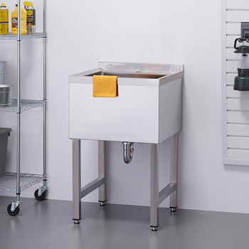 A chrome utility sink with an orange towel draped over its front stands in what appears to be a garage or workshop. To the left, a chrome shelving unit holds buckets and storage bins, while a white slatted wall with various tools and cords hangs on the right.