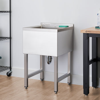 A polished stainless steel utility sink with a high back is shown in a clean, modern laundry room. The sink is on four sturdy legs, with a gray sponge resting on the back ledge, and a rolling black shelf unit with towels and cleaning supplies is to its left.