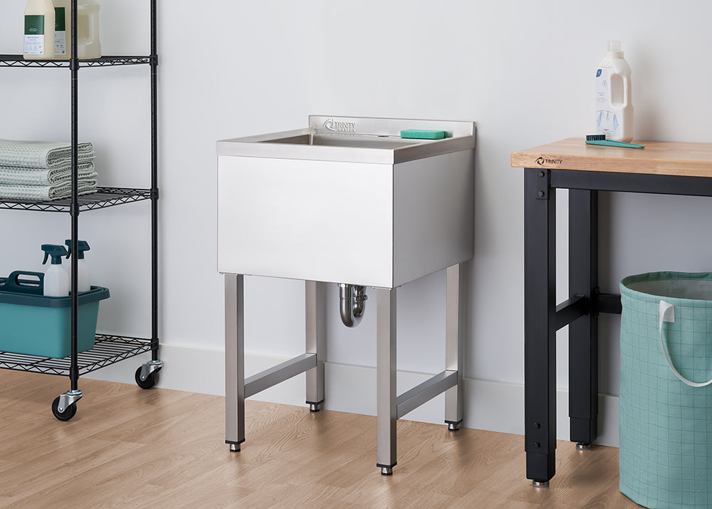 A polished stainless steel utility sink with a high back is shown in a clean, modern laundry room. The sink is on four sturdy legs, with a gray sponge resting on the back ledge, and a rolling black shelf unit with towels and cleaning supplies is to its left.