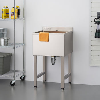 A stainless steel utility sink on four metal legs is centered in a garage or workshop setting. To the left is a chrome wire shelving unit holding various cleaning supplies and storage containers, and to the right is a slatted wall panel with tools and a lantern hanging on it.