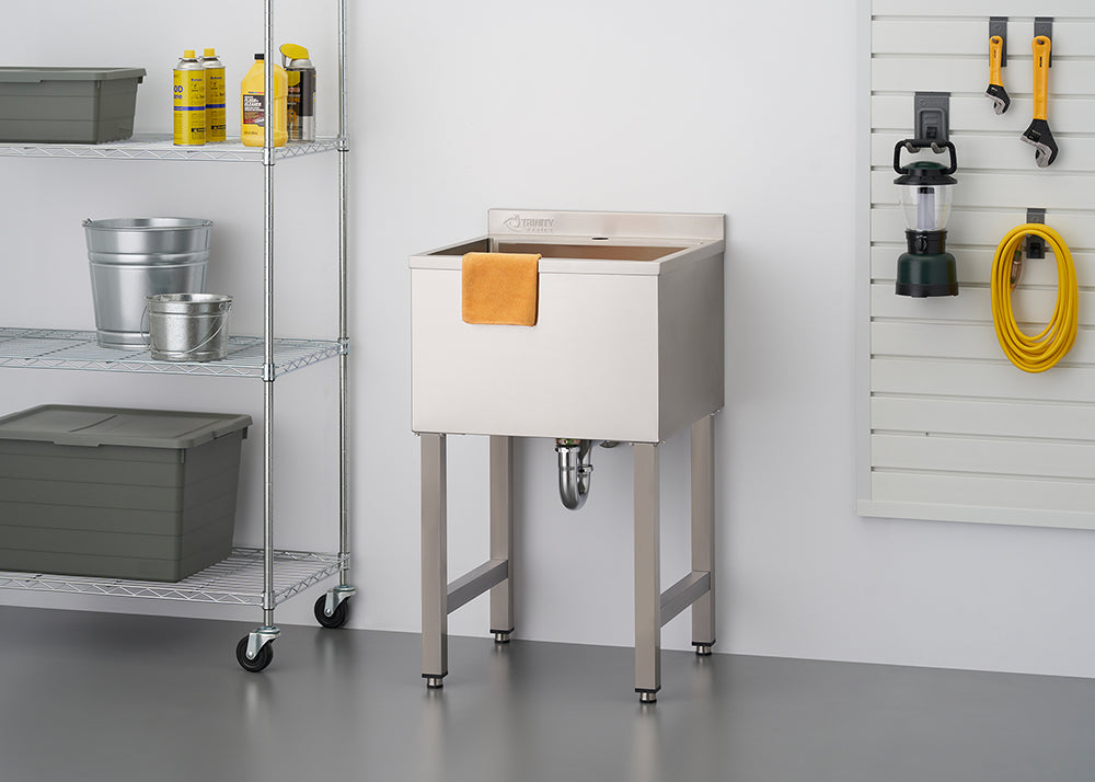 A stainless steel utility sink on four metal legs is centered in a garage or workshop setting. To the left is a chrome wire shelving unit holding various cleaning supplies and storage containers, and to the right is a slatted wall panel with tools and a lantern hanging on it.