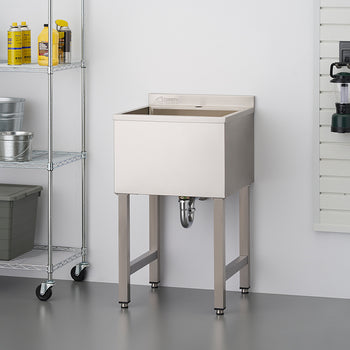 Stainless steel utility sink with legs, situated in a garage or workshop setting next to a wire shelving unit containing various cleaning supplies and storage containers. Tools and a lantern are hung on a slat wall in the background.
