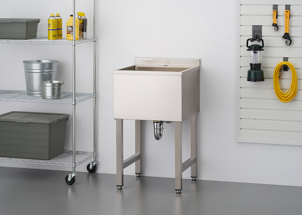 Stainless steel utility sink with legs, situated in a garage or workshop setting next to a wire shelving unit containing various cleaning supplies and storage containers. Tools and a lantern are hung on a slat wall in the background.