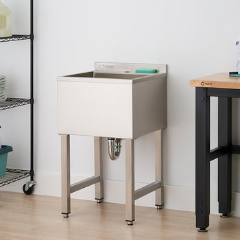 A stainless steel utility sink with legs stands against a white wall in a laundry or utility room. To the left is a black wire shelving unit holding towels and cleaning supplies, and to the right is a wooden workbench next to a green laundry hamper.