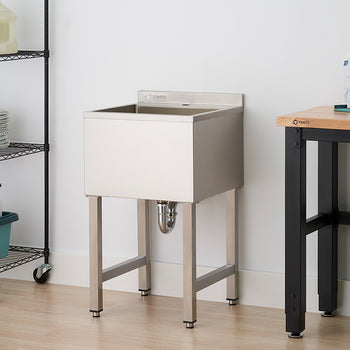 A freestanding, stainless steel utility sink with an attached backsplash is shown in a clean laundry or utility room. The sink stands on four metal legs next to a black wire shelving unit and a wooden-topped workbench.