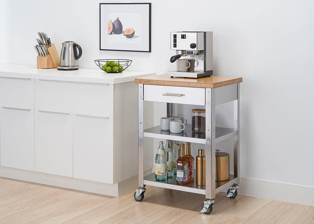 A stainless steel kitchen cart with a light wood top and a white drawer sits on a light-colored floor against a white wall. The cart has two lower shelves holding bottles and mugs, and a coffee maker sits on the top.