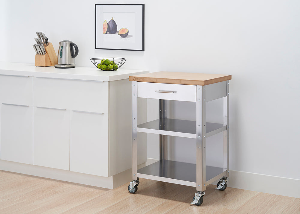 A stainless steel kitchen cart with a light wood top and a drawer is positioned next to a white cabinet. The cart has two shelves and is on wheels, and a framed picture of figs hangs on the wall behind it.