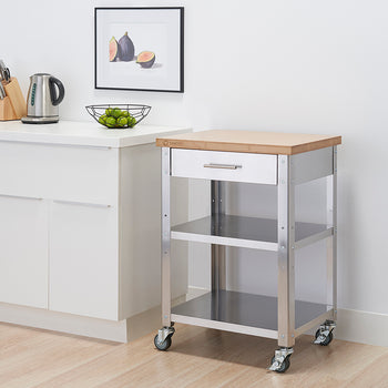 A stainless steel kitchen cart with a light wood top and a drawer is positioned next to a white cabinet. The cart has two shelves and is on wheels, and a framed picture of figs hangs on the wall behind it.