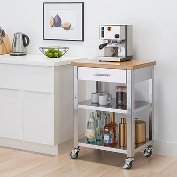 A stainless steel kitchen cart with a light wood top and a white drawer sits on a light-colored floor against a white wall. The cart has two lower shelves holding bottles and mugs, and a coffee maker sits on the top.