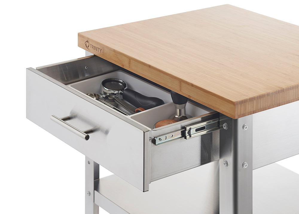 A close-up shot of a stainless steel kitchen cart with a partially extended drawer, revealing organized kitchen utensils inside. The cart features a light-colored bamboo countertop and a visible "TRINITY" logo.