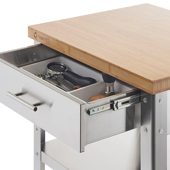 A close-up shot of a stainless steel kitchen cart with a partially extended drawer, revealing organized kitchen utensils inside. The cart features a light-colored bamboo countertop and a visible 