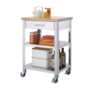 A modern kitchen cart with a stainless steel frame, a wooden top, and four caster wheels. The cart features a drawer, two shelves stocked with kitchen essentials such as jars of lentils, cookbooks, storage containers, and bottles of olive oil.