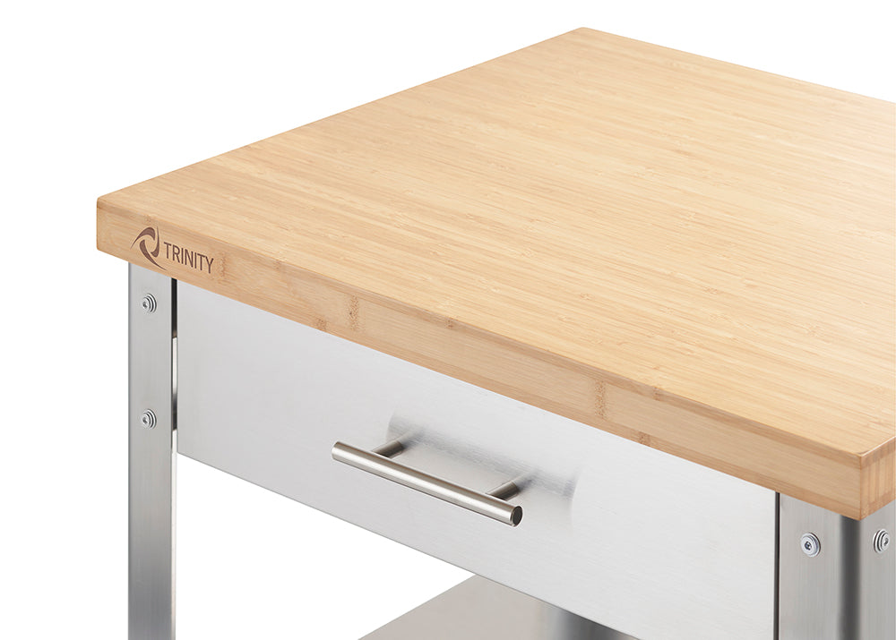 A close-up shot of a light-colored bamboo countertop with the "TRINITY" logo engraved in the corner, atop a stainless steel drawer with a horizontal handle. The cart's frame, also stainless steel, is visible along the edges.