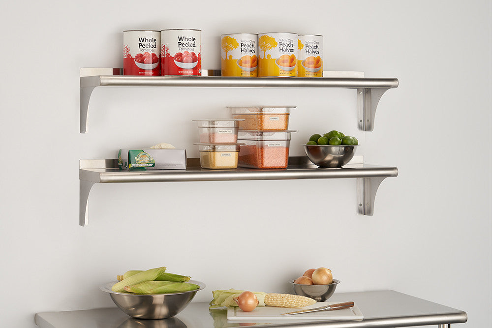 Two stainless steel shelves with bracket supports are mounted on a white wall. The top shelf holds two cans of whole peeled tomatoes and two cans of peach halves, while the bottom shelf holds various food containers and a metal bowl of limes.