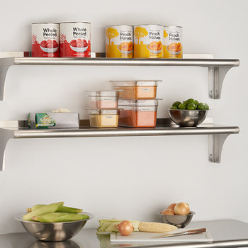 Two stainless steel shelves with bracket supports are mounted on a white wall. The top shelf holds two cans of whole peeled tomatoes and two cans of peach halves, while the bottom shelf holds various food containers and a metal bowl of limes.