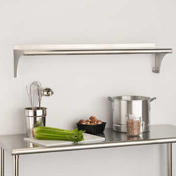 A stainless steel wall-mounted shelf with a towel bar hangs above a matching stainless steel prep table against a white wall. The table holds kitchen items including a container of utensils, celery on a cutting board, a bowl of nuts, a large pot, and a jar of grains.