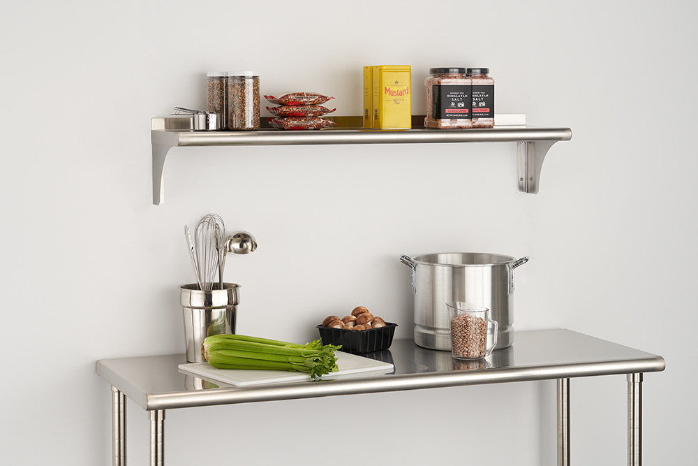 A stainless steel wall shelf is mounted above a stainless steel work table against a white wall. The shelf holds various kitchen items including jars, packages, and a yellow box, while the table below has a celery stalk, a container of nuts, a metal pot, and a utensil holder with whisks.