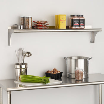 A stainless steel wall shelf is mounted above a stainless steel work table against a white wall. The shelf holds various kitchen items including jars, packages, and a yellow box, while the table below has a celery stalk, a container of nuts, a metal pot, and a utensil holder with whisks.