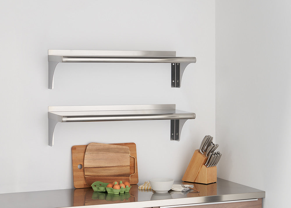 Two stainless steel wall shelves are mounted on a white wall above a stainless steel countertop. The shelves are empty, and the countertop features a wooden cutting board, a carton of eggs, a bowl, and a knife block.
