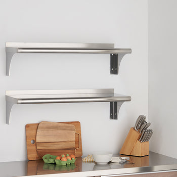 Two stainless steel wall shelves are mounted on a white wall above a stainless steel countertop. The shelves are empty, and the countertop features a wooden cutting board, a carton of eggs, a bowl, and a knife block.
