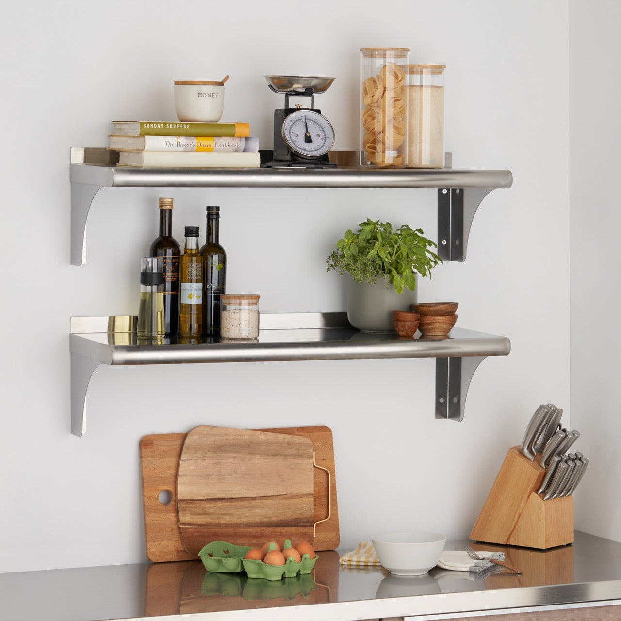 Two stainless steel shelves are mounted on a white wall, holding various kitchen items including books, spices, jars, and a scale. A wooden cutting board and knife block sit on a stainless steel countertop below.