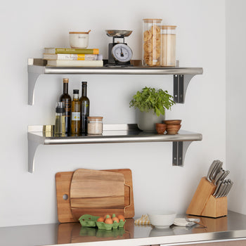 Two stainless steel shelves are mounted on a white wall, holding various kitchen items including books, spices, jars, and a scale. A wooden cutting board and knife block sit on a stainless steel countertop below.