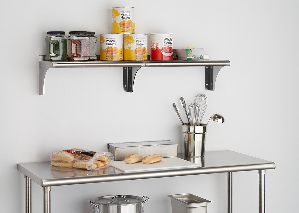 A stainless steel utility table with a shelf above it is shown against a white wall. The table holds a cutting board with two buns, a bag of rolls, and a metal container holding kitchen utensils, while the shelf displays jars of spices and several cans of peaches and tomatoes.