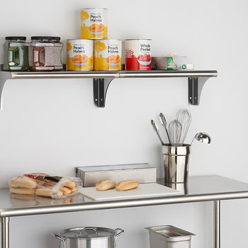 A stainless steel utility table with a shelf above it is shown against a white wall. The table holds a cutting board with two buns, a bag of rolls, and a metal container holding kitchen utensils, while the shelf displays jars of spices and several cans of peaches and tomatoes.