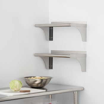 Two stainless steel shelves are mounted on a white wall in a corner, above a stainless steel table with a bowl and a cutting board. The shelves are simple, with a flat surface and a support bracket underneath.