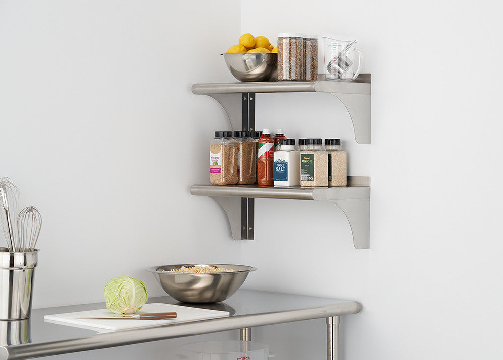 Two stainless steel wall shelves are mounted on a white wall, holding various kitchen items including bowls of lemons, jars, and spice containers. Below the shelves, a stainless steel countertop features a cutting board with a partially chopped cabbage and a metal mixing bowl.