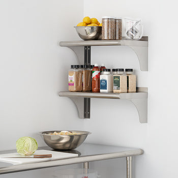 Two stainless steel wall shelves are mounted on a white wall, holding various kitchen items including bowls of lemons, jars, and spice containers. Below the shelves, a stainless steel countertop features a cutting board with a partially chopped cabbage and a metal mixing bowl.